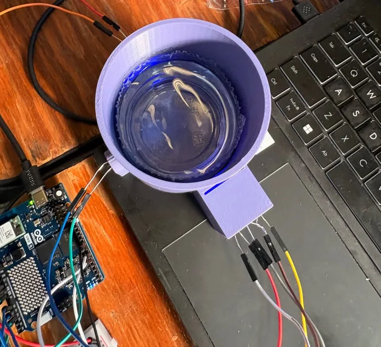 Top view of the station with the lid off and the blue LED on: a clear plastic cup of water inside the purple printed cylinder, blue light at the near wall where the LED box is attached, jumper wires running to the LED legs and to the Arduino UNO Q on the left
