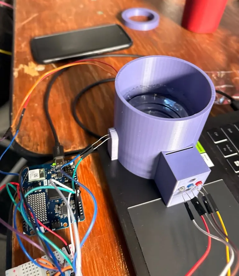 The Pill station on a table: a purple 3D-printed cylinder with a clear cup of water inside, the purple LED box on its side with white, blue and red LEDs wired in, a photoresistor holder on the left wall, and an Arduino UNO Q with jumper wires in front